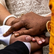 Multiple hands of different skin tones stacked together in a gesture of connection and support, adorned with traditional bracelets and malas, against a patterned fabric background