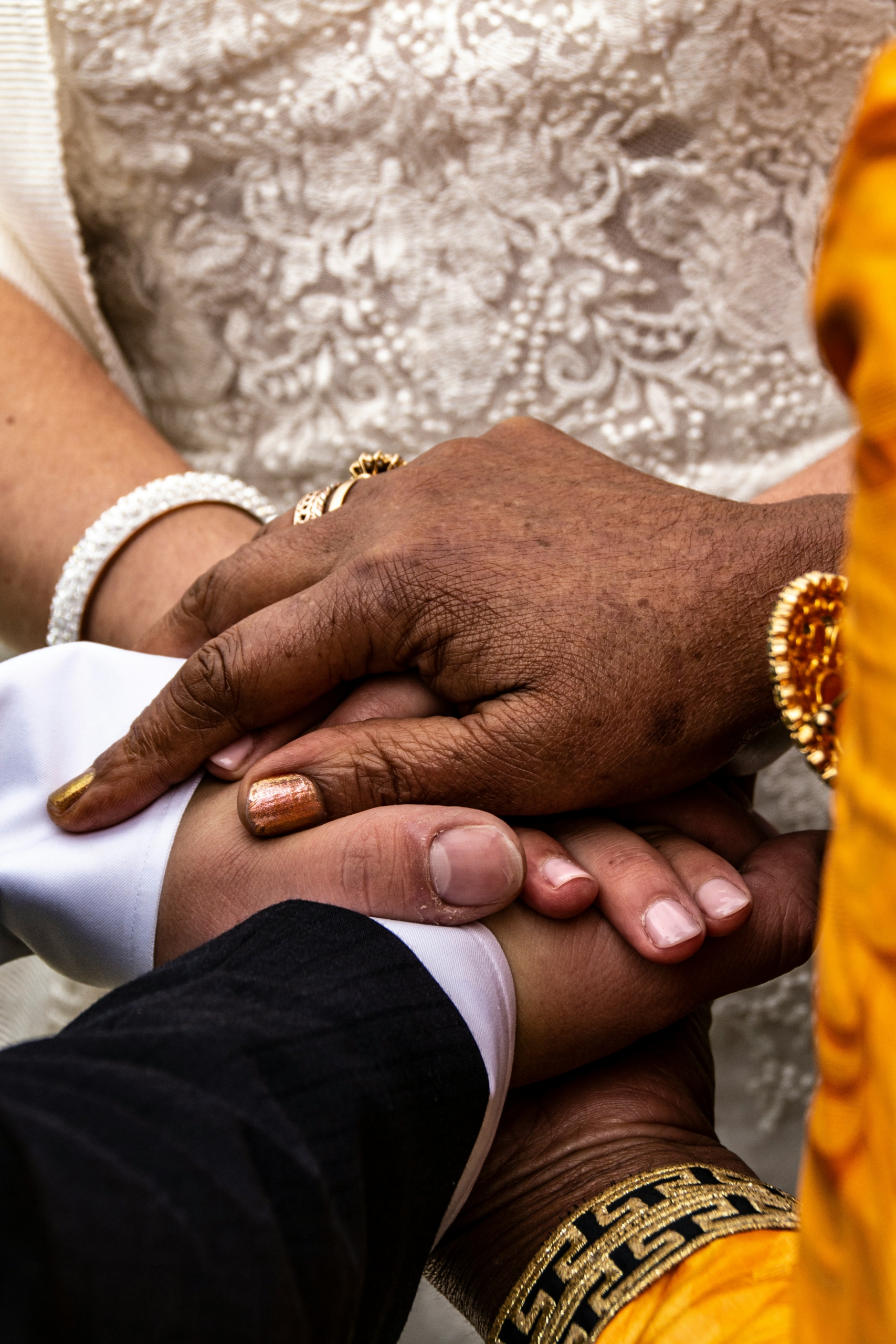 Multiple hands of different skin tones stacked together in a gesture of connection and support, adorned with traditional bracelets and malas, against a patterned fabric background