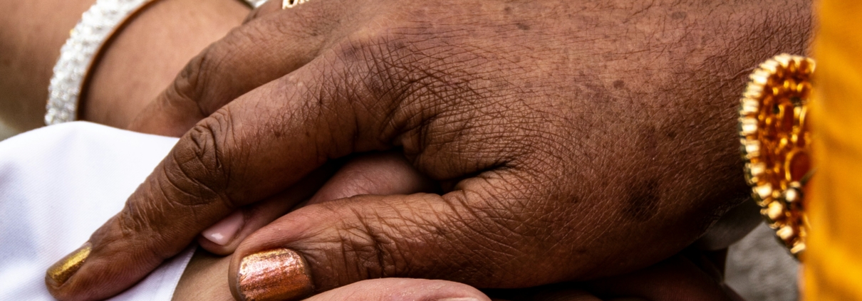 Multiple hands of different skin tones stacked together in a gesture of connection and support, adorned with traditional bracelets and malas, against a patterned fabric background