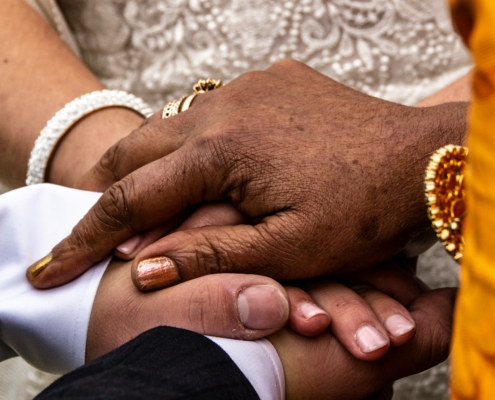 Multiple hands of different skin tones stacked together in a gesture of connection and support, adorned with traditional bracelets and malas, against a patterned fabric background