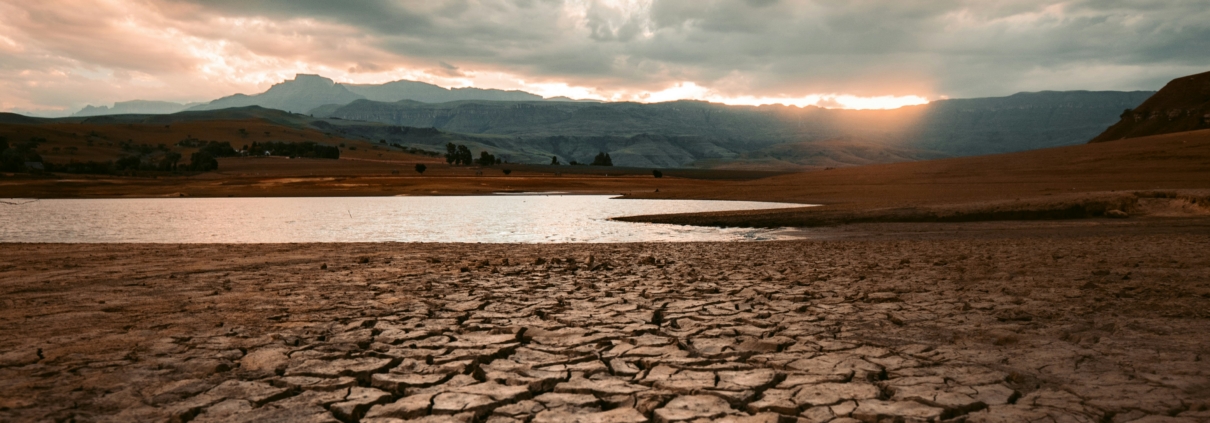 Cracked dry earth in the foreground leading toward a still body of water, with dramatic storm clouds and breaking light in the sky above a mountain range at sunset.