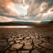 Cracked dry earth in the foreground leading toward a still body of water, with dramatic storm clouds and breaking light in the sky above a mountain range at sunset.