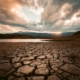 Cracked dry earth in the foreground leading toward a still body of water, with dramatic storm clouds and breaking light in the sky above a mountain range at sunset.