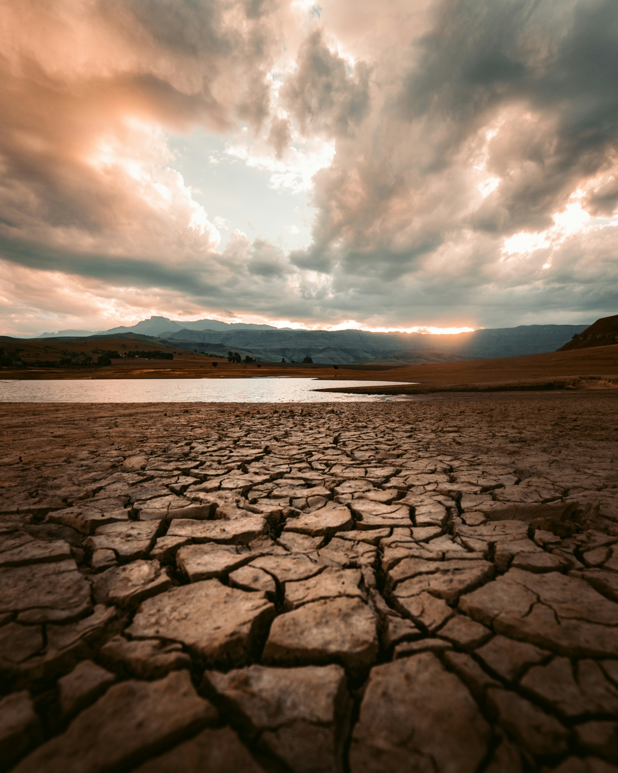 Cracked dry earth in the foreground leading toward a still body of water, with dramatic storm clouds and breaking light in the sky above a mountain range at sunset.